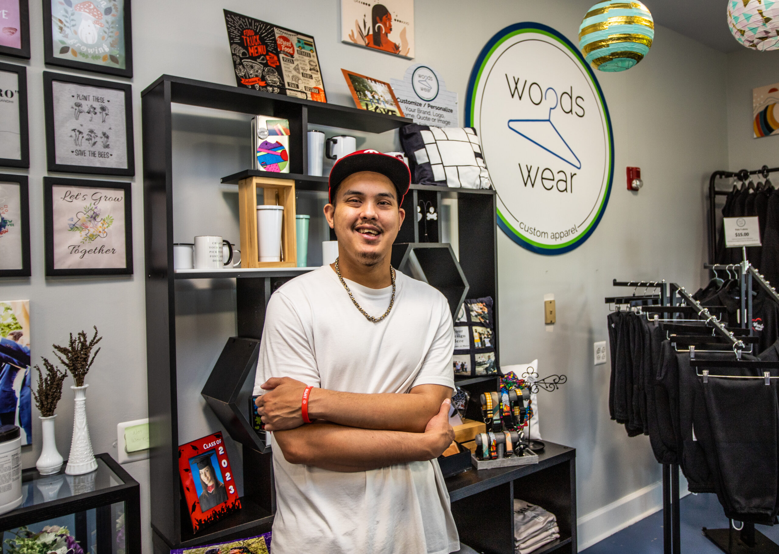 mans stands smiling with arms folded in front of a retail shelf near a sign that says "woods wear customer apparel" with a clothes hanger icon