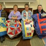 four women sitting in front of a fireplace holding custom quilts